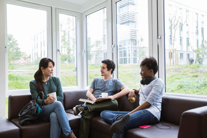Three students sat drinking coffee and talking, one is holding a book.